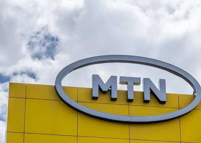 A close-up photograph of an MTN corporate sign mounted on a bright yellow tiled wall. The 3D silver 'MTN' lettering is enclosed in an oval frame against a background of a blue sky with white clouds.