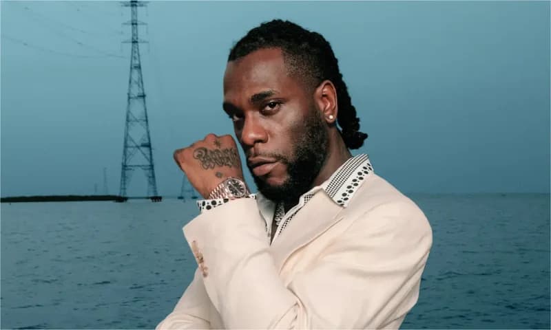 A professional portrait of Nigerian Afrobeats star Burna Boy wearing a cream blazer and a luxury wristwatch. He is posing in front of a body of water with a large electricity transmission tower visible in the background under a blue sky.