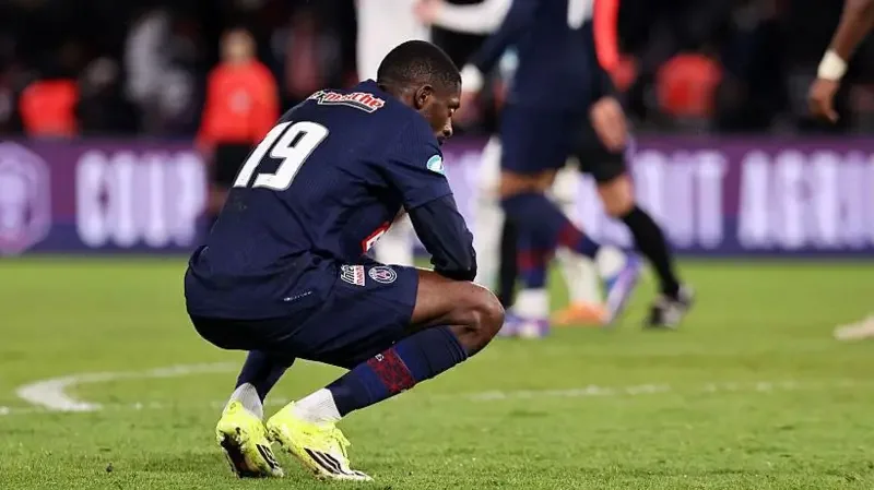 A dejected Paris Saint-Germain player wearing the number 19 jersey squats on the pitch following a match. The scene captures the emotional impact of a defeat, likely after the team's exit from the Coupe de France as indicated by the background branding.