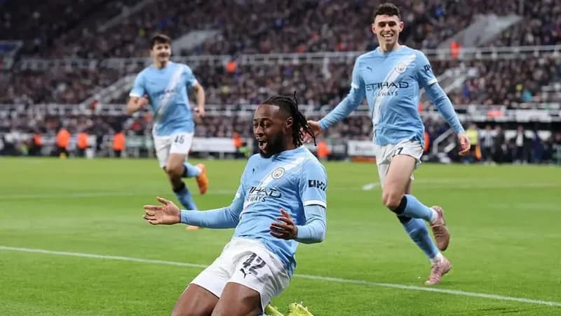 Manchester City players celebrate a goal during a football match against Newcastle United. The foreground shows Oscar Bobb kneeling on the pitch with arms outstretched in celebration, while teammates Phil Foden and others run toward him in front of a crowded stadium.