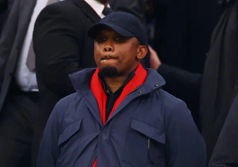 The image shows Samuel Eto'o, seemingly at a public event or match. He is wearing a dark cap, a blue jacket, and a red scarf, looking slightly off-center with a contemplative expression. Other people are visible in the background, suggesting a crowded environment.
