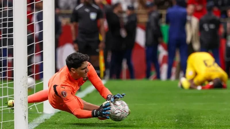 The image captures a moment from a football match, likely a penalty shootout, with a goalkeeper in an orange uniform diving to save the ball. Another player in a yellow uniform is seen on the ground in the background, possibly reacting to the save or miss. The image conveys the tension and drama of a critical moment in a sporting event.