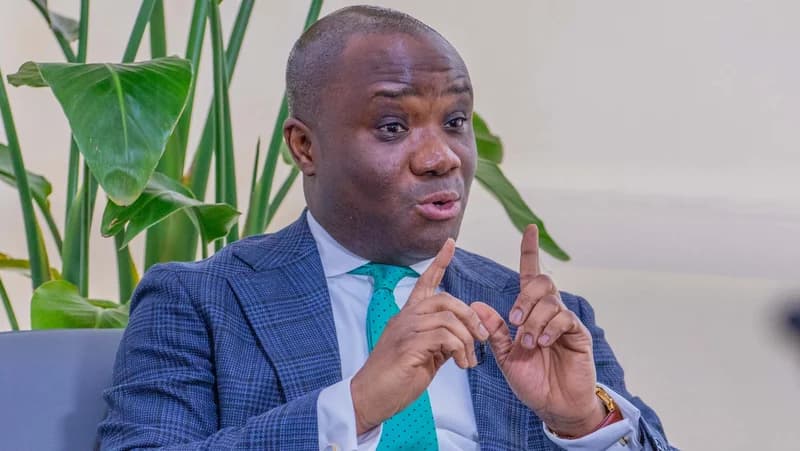 Felix Kwakye Ofosu, a prominent Ghanaian politician, is captured mid-speech during an interview or official event. He is dressed in a blue checkered suit and a green tie, gesturing with his hands to emphasize his point against a backdrop of indoor greenery.
