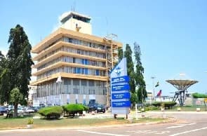 The image features the Ghana Civil Aviation Authority (GCAA) headquarters building in Accra, characterized by its multi-story structure and an integrated control tower. A prominent blue directional sign stands in the foreground on a landscaped lawn, with a large satellite communication dish visible in the background under a clear sky.