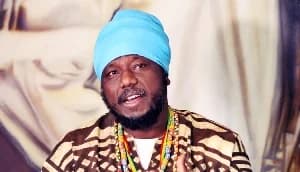 A close-up portrait of Ghanaian reggae musician and radio presenter Blakk Rasta. He is wearing a distinctive light blue turban and a brown and cream patterned garment with colorful beads around his neck.