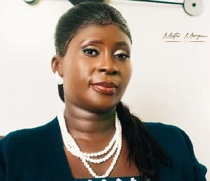 A medium shot depicts Esther Smith, who is defending Pastor Elvis Agyemang in an extortion case. She is wearing a dark blue jacket over a light-colored top and a multi-strand pearl necklace. The background is a plain white wall with a modern clock visible in the upper left corner.