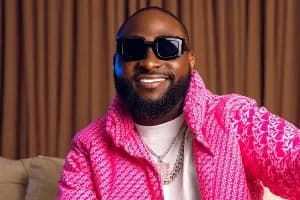 A portrait of Nigerian Afrobeats star Davido smiling while wearing dark sunglasses and a vibrant pink textured jacket. He is adorned with multiple silver chains over a white t-shirt, set against a background of brown pleated curtains.