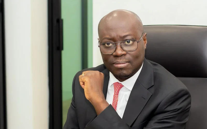 A portrait of Ghanaian politician Cassiel Ato Forson, seen wearing a dark suit, white shirt, and a red patterned tie. He is seated in a black office chair and looking directly at the camera with a serious and professional expression.
