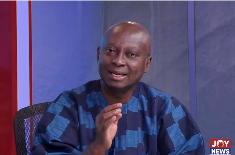 A photograph of Ghanaian politician Samuel Atta Akyea speaking during a media appearance. He is wearing a traditional blue and navy checkered smock and gesturing with his right hand while seated in a studio setting with a Joy News logo visible.