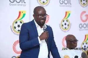 Kurt Okraku, the President of the Ghana Football Association (GFA), is pictured speaking into a microphone at an official event. He is wearing a blue blazer over a white shirt and glasses, standing in front of a backdrop featuring the GFA logo and GOIL branding. A second individual is partially visible in the background seated to the right.