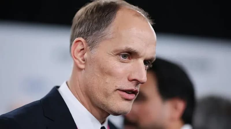 A close-up photograph of football manager Thomas Tuchel looking off-camera with a serious expression. He is dressed in a professional suit jacket and white shirt, set against a softly blurred background typical of a media event.