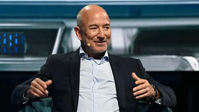 This is a photo of Jeff Bezos sitting on a chair. He is wearing a blue shirt, a black suit jacket, and a microphone, and his hands are gesturing outwards. The photo is relevant to the article because it mentions Jeff Bezos' Blue Origin.