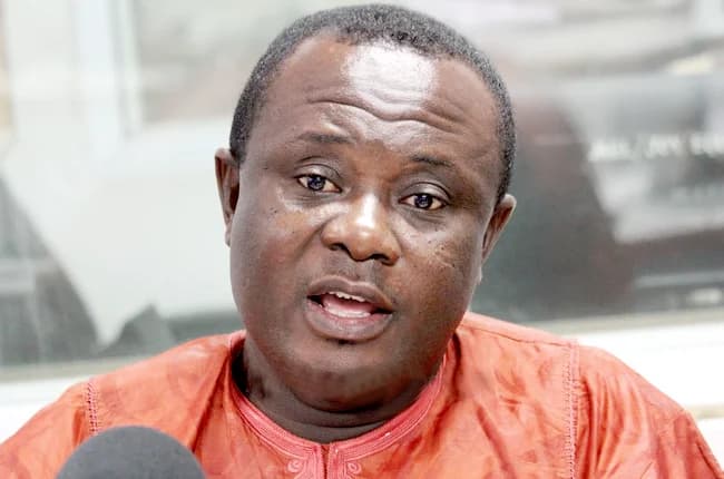This is a close-up shot of Osei Owusu, likely speaking at an event or giving an interview. He is wearing an orange, patterned shirt and appears to be in a room with some other people or equipment in the background. The photo is well-lit and focused.