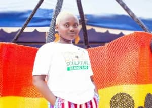 This image shows Juliana Gharbin standing in front of what appears to be the beads sculpture. She is wearing a white t-shirt with text related to the sculpture. The backdrop is partially visible and appears to be made of beads in varying colors.