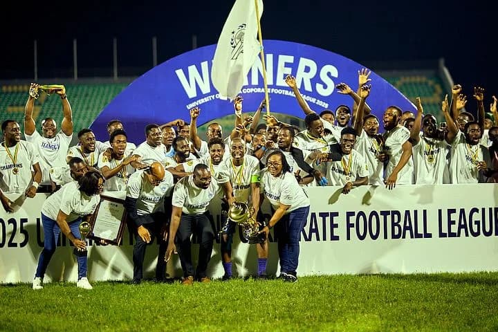Members of the Ghana Civil Aviation Authority (GCAA) football team celebrate their victory at the University of Ghana Corporate Football League 2025. The group is gathered on a pitch under a blue 'WINNERS' arch, hoisting a gold trophy, a framed certificate, and a white flag while wearing 'TEAM GCAA' branded shirts and medals.