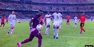 Ghanaian goalkeeper Richard Ofori is captured touching the ball as it rolls out of play during a crucial football match. Several players in the white Ghana national team kit and the red opponent kit are visible on the pitch within a packed stadium.