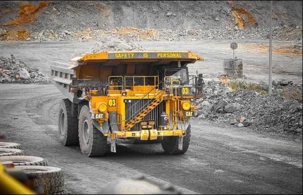 A large yellow Caterpillar mining dump truck carries a heavy load of rocks through an open-pit mine. The vehicle features the slogan 'SAFETY IS PERSONAL' prominently on its front visor, set against a backdrop of excavated earth and stone.