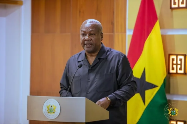 President John Dramani Mahama is depicted delivering a formal address from behind a lectern. He is positioned in front of the flag of Ghana and a wood-paneled wall, wearing a dark button-down shirt.