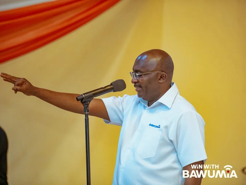 Dr. Mahamudu Bawumia is pictured speaking at a political event, wearing a light blue branded shirt and spectacles. He has his right arm outstretched with three fingers raised, while standing behind a microphone against a yellow background with red drapery.