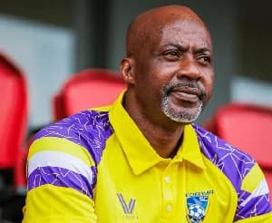 A close-up portrait of Ibrahim Tanko, the coach of Medeama SC, wearing the team's distinctive yellow and purple jersey. He is shown with a grey-flecked beard and a thoughtful expression, seated in front of empty red stadium seats.