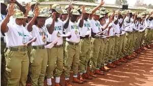A line of National Youth Service Corps (NYSC) members in Nigeria are standing outdoors during an official ceremony. They are dressed in their signature white t-shirts with 'NYSC' branding and khaki trousers, with many raising their right hands as they appear to be taking an oath or reciting a pledge.
