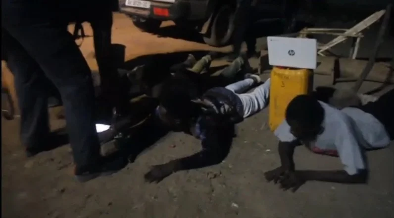 A night-time scene showing three suspects lying face-down on the ground during a police operation in Ghana. Law enforcement officers are seen standing over the individuals, with seized items such as a white HP laptop and a yellow jerrycan visible nearby.