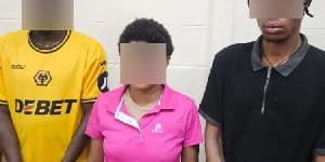 Three individuals, two males and one female, are standing against a light-colored wall with their faces blurred for legal reasons. They are dressed in casual clothing, including a yellow Wolverhampton Wanderers jersey and a pink polo shirt, appearing to be in police custody.