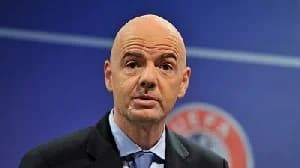 A close-up portrait of FIFA President Gianni Infantino speaking at an event. He is wearing a dark suit jacket, a light blue shirt, and a blue tie, positioned against a blue backdrop featuring a blurred UEFA logo.