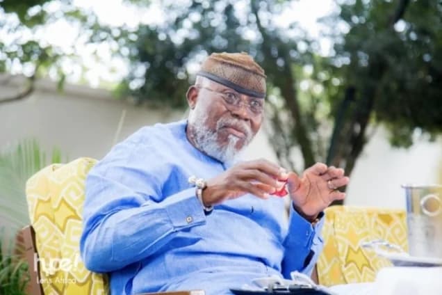 A portrait of Dr. Nyaho Nyaho-Tamakloe, a prominent Ghanaian politician and founding member of the NPP, seated outdoors in a blue traditional outfit. He is captured gesturing with his hands while speaking, set against a background of lush green foliage.