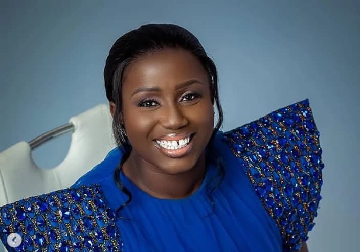 A studio portrait of Ghanaian gospel singer Diana Hamilton, who is smiling warmly at the camera. She wears a striking royal blue gown featuring dramatic, jewel-encrusted shoulder pads against a muted blue background.