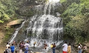 A scenic view of a multi-tiered waterfall in a lush forest setting, with numerous people gathered at the base for recreation. Tourists are seen swimming, wading, and standing on the rocks while enjoying the natural attraction, which appears to be a popular Ghanaian destination like Boti Falls.