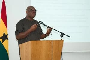 Yofi Grant, the CEO of the Ghana Investment Promotion Centre (GIPC), is pictured speaking at a wooden podium during a business presentation. He is wearing a dark traditional tunic and glasses, standing next to the Ghanaian national flag with a projector screen visible in the background.