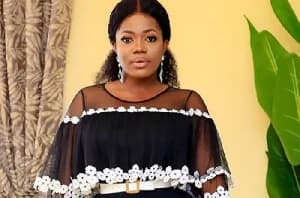 A portrait of Ghanaian musician Mzbel, seen wearing a black mesh dress with white floral lace trimmings and a white belt. She is standing indoors in front of a yellow wall with a patterned curtain and a green leaf visible in the background.
