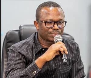 A close-up photograph of Kofi Ofosu Nkansah, the CEO of the National Entrepreneurship and Innovation Programme (NEIP), wearing black-rimmed glasses and a black pinstriped shirt. He is speaking into a microphone while seated in a black executive chair, likely during a formal board meeting or business presentation.