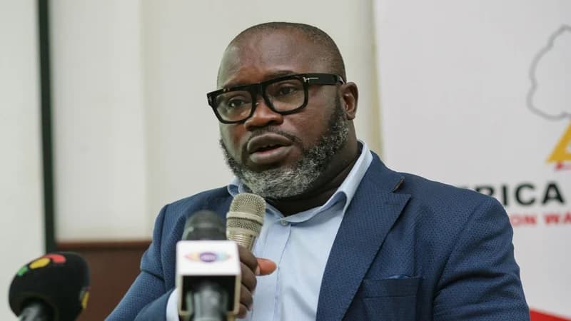 A close-up photograph of Kofi Asare, Executive Director of Africa Education Watch (Eduwatch), speaking at an event. He is wearing thick-rimmed black glasses and a blue blazer while addressing microphones, with a partial Eduwatch banner visible in the background.
