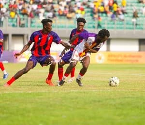 Action shot from a Ghana Premier League match showing a Hearts of Oak player in a white and gold kit controlling the ball. He is being closely marked by two Eleven Wonders players wearing their signature purple and red striped jerseys in front of a stadium crowd.