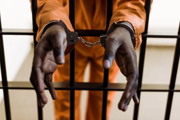 A close-up photograph showing a person's hands in metal handcuffs, positioned through the vertical bars of a jail cell. The individual is wearing a bright orange jumpsuit, which is a common visual representation of incarceration or being in police custody.