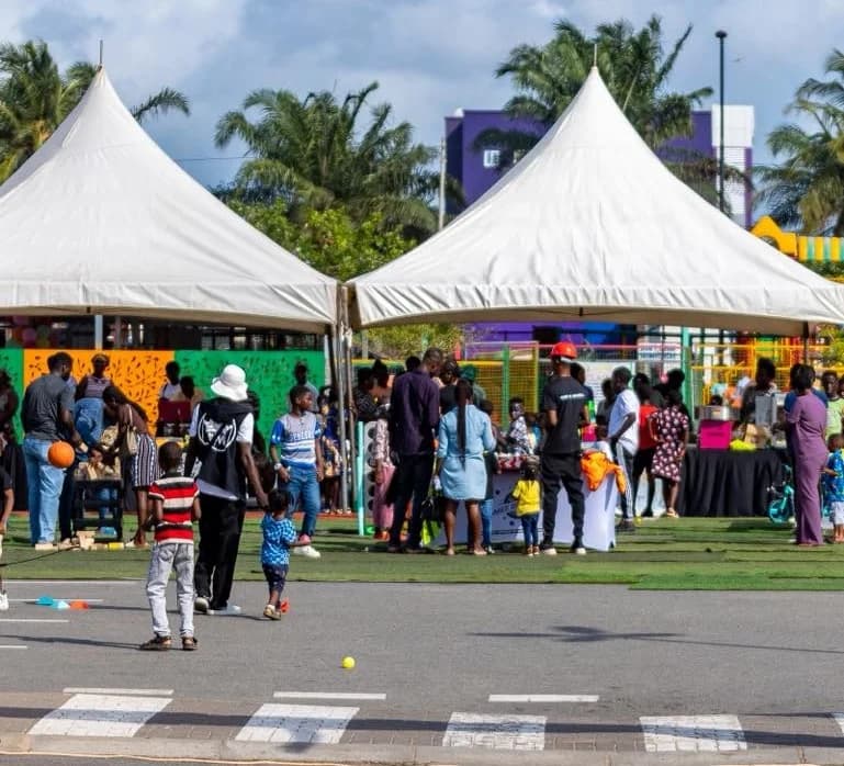 A lively outdoor scene at the Achimota Retail Centre featuring large white peaked tents set up on artificial turf. A diverse crowd of adults and children are gathered around vendor stalls and activity areas during the 'Joy at the Mall' climax event.