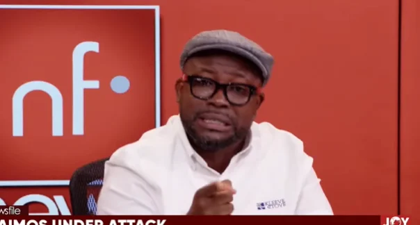 Senyo Hosi is captured during a televised appearance on Joy News, wearing a white shirt, a grey flat cap, and black glasses. He appears to be making a point emphatically with a raised hand against a red studio backdrop featuring the 'Newsfile' logo.