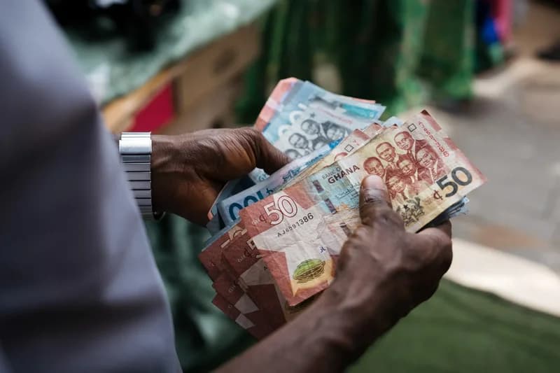 A close-up photograph shows a person counting Ghanaian Cedi banknotes. The person is holding a mix of denominations, including fifty and one hundred cedi notes, which feature the portraits of the 'Big Six' and the cocoa pod symbol.