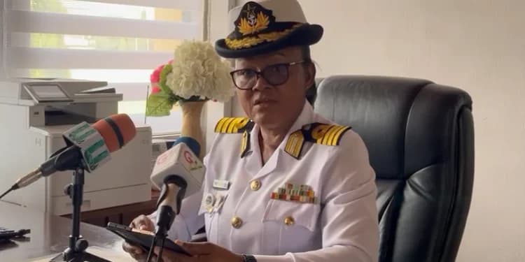 A high-ranking female officer of the Ghana Navy is pictured seated at a desk, addressing the press with several microphones positioned in front of her. She is wearing a formal white naval uniform and a peaked cap, representing the Ghana Armed Forces during an official statement.