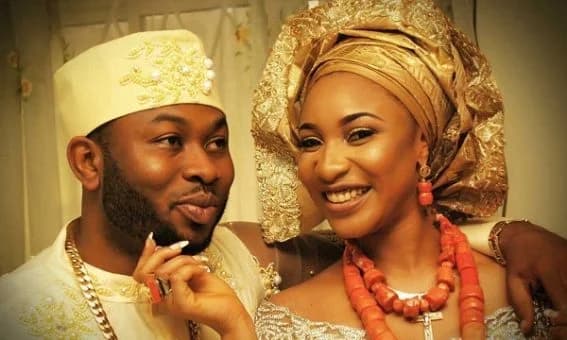 A photograph of Nigerian actress Tonto Dikeh and her ex-husband, Olakunle Churchill, in traditional West African wedding attire. Both are smiling warmly as the woman wears an ornate gold headtie (gele) with coral beads, while the man wears a matching cream-colored cap and gold accessories.