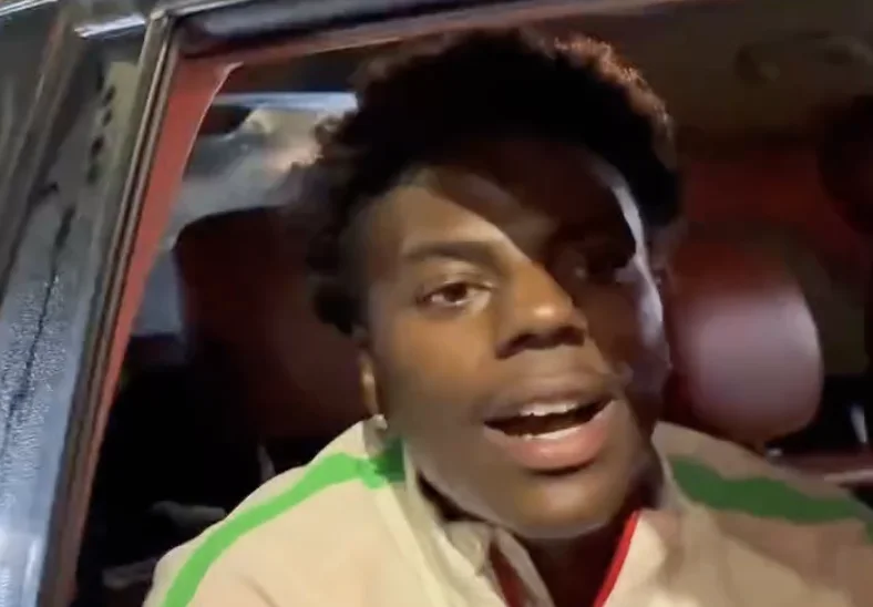 A close-up photograph of YouTuber iShowSpeed sitting inside a vehicle during his arrival in Ghana. He is wearing a light-colored tracksuit with green stripes and appears to be speaking or reacting excitedly to something outside the car.