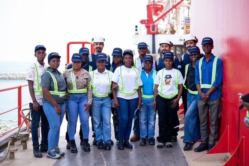 A group of Takoradi Technical University (TTU) engineering students and officials pose for a photograph on the deck of a powership. They are wearing industrial safety gear, including high-visibility vests and branded caps, with the ship's red hull and the ocean visible in the background.