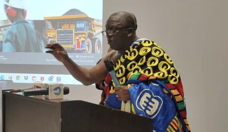 Nana Nketsia V, a prominent Ghanaian traditional leader, is captured speaking at a podium during a business-related presentation. He is dressed in traditional cloth featuring Adinkra symbols, gesturing for emphasis while holding a microphone against a backdrop of a screen showing mining operations.