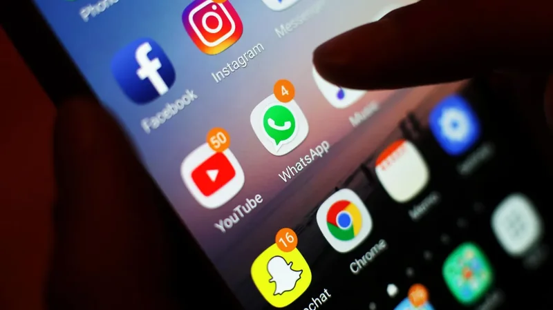 A close-up photograph of a smartphone screen displaying several popular social media app icons including Facebook, Instagram, WhatsApp, and YouTube. A finger is shown interacting with the device, which features several red notification badges indicating unread alerts across different platforms.