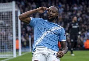 A professional action photograph of Ghanaian footballer Antoine Semenyo celebrating on a football pitch. He is seen wearing a light blue jersey, performing a salute-style gesture with his hand to his forehead, while a goalpost and a blurred stadium crowd are visible in the background.