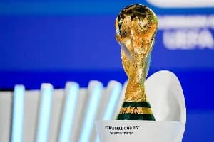 The iconic gold FIFA World Cup trophy is displayed prominently against a vibrant blue background. It sits atop a white stand that contains text identifying it as the official winner's trophy for the 2026 tournament.