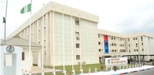 The image shows the exterior of the Defence Headquarters building in Nigeria, a large multi-story cream-colored structure. A Nigerian green and white flag is seen flying on a pole in the foreground next to a prominent sign identifying the facility.