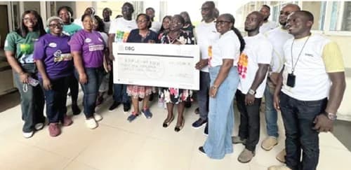 A group of Development Bank Ghana (DBG) employees are gathered for a charitable presentation. They are holding a large ceremonial check addressed to the Korle-Bu NICU for the sum of GHC 39,800. The photo captures a corporate social responsibility event where staff are seen giving back to the local healthcare community.